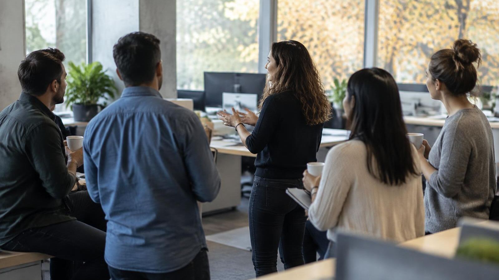 A manager leads an informal team stand-up in an open office with notebooks and coffee cups in soft early fall light